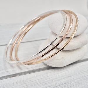 Puede incluir: Tres brazaletes de metal martillado en tonos plateado, oro rosa y dorado descansan sobre una pila de piedras blancas lisas. Los brazaletes tienen una superficie texturizada y un diseño simple y elegante, ideal para el uso diario.