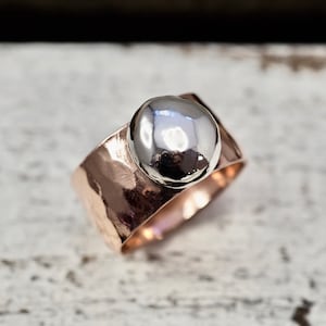 以下が含まれることがあります： ハンマーで叩かれたローズゴールドのバンドリング。上面には滑らかなシルバー調のドームが乗っています。