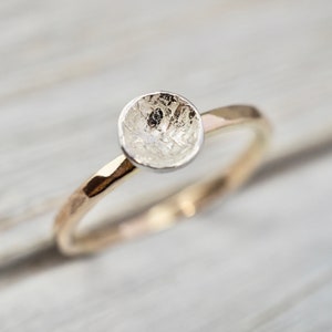 May include: A gold ring with a textured, silver-coloured setting. The ring has a hammered band and a round, cup-like setting. The ring is set against a light-coloured background.
