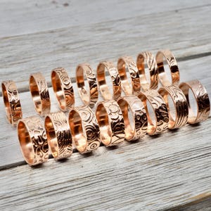 May include: A collection of 16 rose gold rings with various embossed designs. The rings are arranged in a row on a light wood surface.