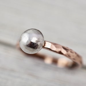 May include: A silver ball ring with a hammered rose gold band. The ring features a shiny, spherical silver bead as its focal point, set atop a textured, rose gold band. The band has a hammered finish.