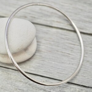 May include: A simple silver bangle bracelet, made of a single, smooth, round piece of metal. The bracelet is lying on a light wood surface.