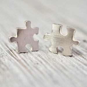 Puede incluir: Un par de pendientes de plata con forma de pieza de puzzle. Los pendientes son pequeños y delicados, y son perfectos para añadir un toque de fantasía a cualquier atuendo.