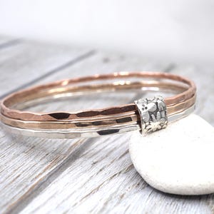 May include: Three hammered metal bangles in rose gold, silver, and gold tones, held together by a decorative silver clasp. The bangles are displayed on a white stone against a light wood background. The jewellery is simple and elegant.