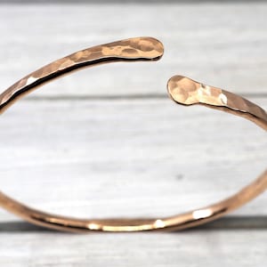 May include: A rose gold-toned open cuff bracelet featuring a hammered, textured surface. The bracelet has a circular band that tapers slightly at the two rounded, open ends, creating a continuous, reflective metallic finish throughout the piece.