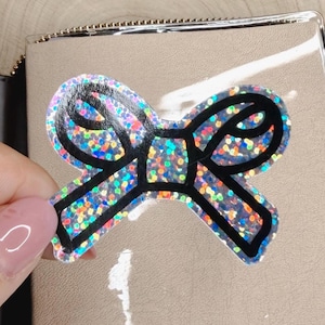Può includere: Un adesivo a forma di fiocco olografico con un bordo nero spesso. L'adesivo presenta una superficie multicolore, riflettente e scintillante che cattura la luce, creando un effetto luccicante. È applicato sulla copertina in plastica trasparente di un taccuino.