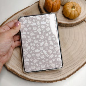 Può includere: Una custodia protettiva trasparente per tablet con un motivo ripetuto di piccoli volti di orso bianchi, nuvole e fiori a quattro petali su uno sfondo color tortora chiaro. La custodia presenta angoli rinforzati e una finitura liscia e lucida.