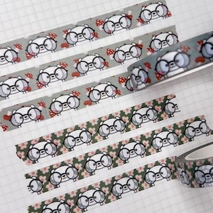 Può includere: Due rotoli di nastro adesivo decorativo con un motivo ripetuto di orsi bianchi dei cartoni animati che indossano occhiali neri e piccoli funghi rossi. Il nastro è mostrato su una pagina di agenda a quadretti con un calendario di luglio.