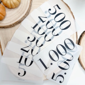 Può includere: Una collezione di carte rettangolari traslucide con numeri stampati in nero, tra cui 500, 200, 100, 50, 20, 10, 5, 1.000 e 5.000. Ogni carta riporta il testo AUBRIEBOUTIQUE in piccole lettere nere nella parte inferiore del design.