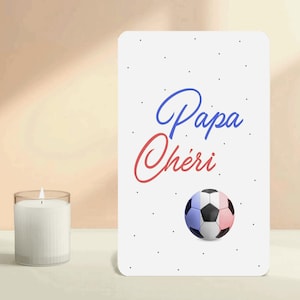 Pode incluir: Um cartão retangular branco com as palavras "Papa Chéri" em escrita azul e vermelha, e uma bola de futebol com as cores da bandeira francesa. Pequenos pontos pretos estão espalhados pelo cartão. Uma vela branca num castiçal transparente à esquerda.