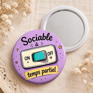 Puede incluir: Un espejo compacto redondo morado con las palabras "Sociable à temps partiel" y un diseño de interruptor "ON/OFF". El espejo está abierto, revelando una superficie reflectante. El diseño incluye estrellas y corazones.