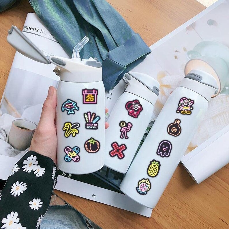 May include: Three white water bottles with colorful neon stickers. The stickers feature various designs, including a calendar, a palm tree, a flamingo, a ghost, a pineapple, and a dartboard.