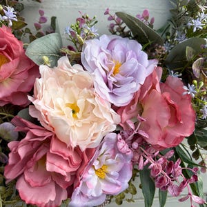 May include: A close-up of a floral wreath featuring large, multi-toned faux peonies in shades of pink, peach, and lavender. Green leaves and small purple and blue flowers are interspersed throughout the arrangement, creating a vibrant and textured display.