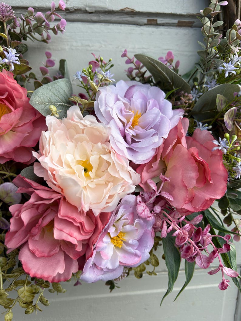 May include: A close-up of a floral wreath featuring large, multi-toned faux peonies in shades of pink, peach, and lavender. Green leaves and small purple and blue flowers are interspersed throughout the arrangement, creating a vibrant and textured display.