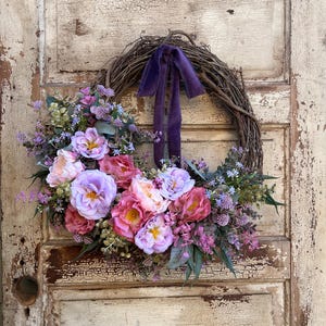 May include: A floral wreath with a grapevine base, adorned with pink, purple, and lavender flowers, and greenery. A deep purple ribbon is tied at the top. The wreath is displayed on a distressed, off-white wooden door.