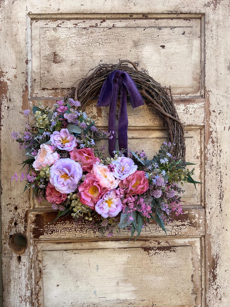 May include: A floral wreath with a grapevine base, adorned with pink, purple, and lavender flowers, and greenery. A deep purple ribbon is tied at the top. The wreath is displayed on a distressed, off-white wooden door.