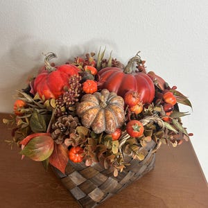 May include: A fall-themed arrangement in a woven basket. It features vibrant orange and red pumpkins of various sizes, interspersed with faux foliage, berries, and pinecones. The basket is a dark brown with a woven pattern, creating a rustic aesthetic.