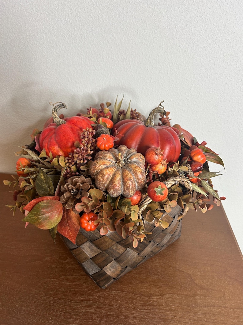 May include: A fall-themed arrangement in a woven basket. It features vibrant orange and red pumpkins of various sizes, interspersed with faux foliage, berries, and pinecones. The basket is a dark brown with a woven pattern, creating a rustic aesthetic.