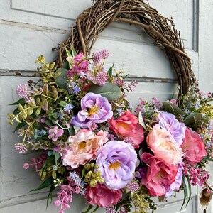 May include: A decorative wreath featuring a natural grapevine base adorned with an array of colorful faux flowers. The arrangement includes large, vibrant blooms in shades of pink, purple, and peach, complemented by green foliage.
