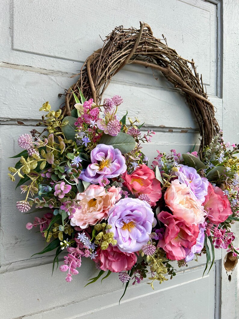 May include: A decorative wreath featuring a natural grapevine base adorned with an array of colorful faux flowers. The arrangement includes large, vibrant blooms in shades of pink, purple, and peach, complemented by green foliage.