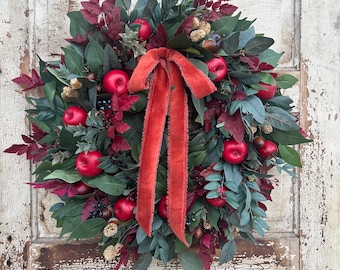 May include: A circular wreath featuring deep green foliage, dark red leaves, and bright red artificial apples. A long, frayed-edge velvet ribbon in a burnt orange hue is tied in a bow at the top center, with two long tails hanging down the middle.