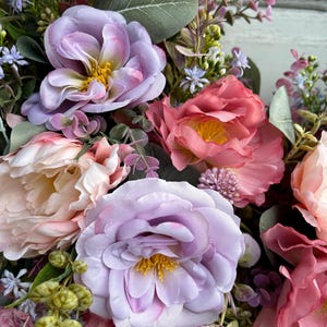 May include: Close-up of a floral arrangement featuring large, soft-petaled flowers in shades of lavender, pink, and peach. The arrangement includes greenery and smaller purple and white blossoms, creating a full and textured display. The flowers have yellow centers.