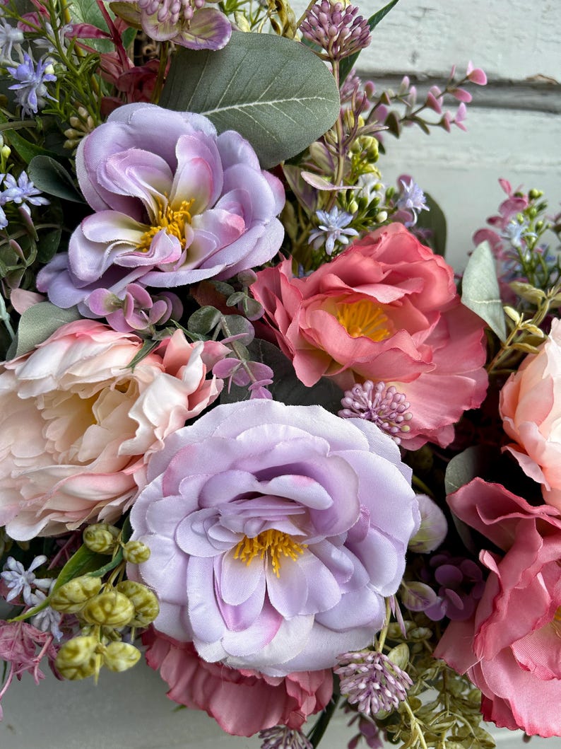 May include: Close-up of a floral arrangement featuring large, soft-petaled flowers in shades of lavender, pink, and peach. The arrangement includes greenery and smaller purple and white blossoms, creating a full and textured display. The flowers have yellow centers.
