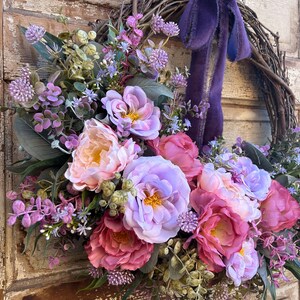 May include: A floral wreath featuring a variety of faux flowers in shades of pink, purple, and peach. The wreath is made of woven branches and has a purple ribbon. The flowers include peonies and other blooms.