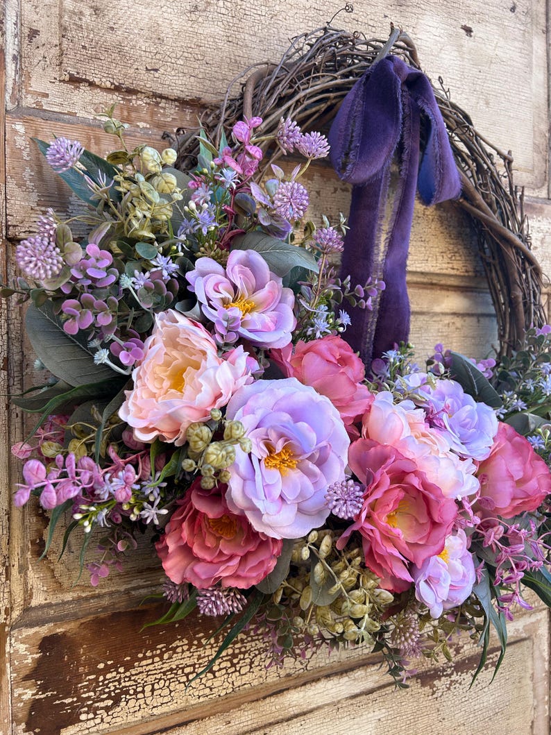 May include: A floral wreath featuring a variety of faux flowers in shades of pink, purple, and peach. The wreath is made of woven branches and has a purple ribbon. The flowers include peonies and other blooms.