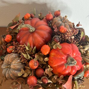 May include: A fall-themed centerpiece featuring several orange pumpkins of varying sizes, surrounded by faux foliage, berries, and pinecones. The arrangement includes a mix of textures and colors, creating a warm and inviting autumn display.