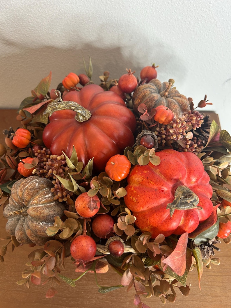 May include: A fall-themed centerpiece featuring several orange pumpkins of varying sizes, surrounded by faux foliage, berries, and pinecones. The arrangement includes a mix of textures and colors, creating a warm and inviting autumn display.