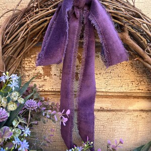 May include: A grapevine wreath adorned with purple faux flowers and greenery. A long, fringed, purple velvet ribbon is tied to the top of the wreath. The background is a distressed, cream-colored wooden surface.