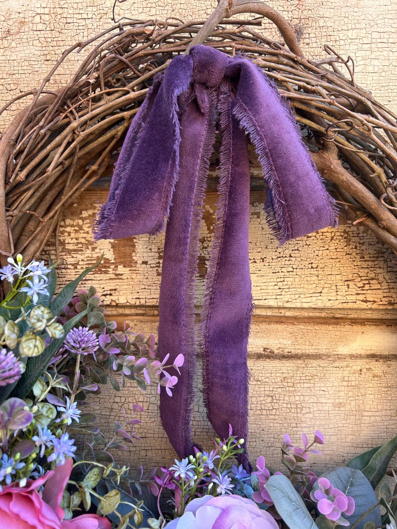May include: A grapevine wreath adorned with purple faux flowers and greenery. A long, fringed, purple velvet ribbon is tied to the top of the wreath. The background is a distressed, cream-colored wooden surface.