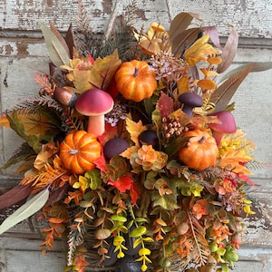 May include: A fall-themed hanging basket overflowing with faux pumpkins, mushrooms, and colorful leaves in shades of orange, red, and yellow. The basket is made of woven twigs and is suspended by a rope.