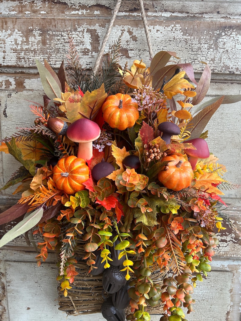 May include: A fall-themed hanging basket overflowing with faux pumpkins, mushrooms, and colorful leaves in shades of orange, red, and yellow. The basket is made of woven twigs and is suspended by a rope.