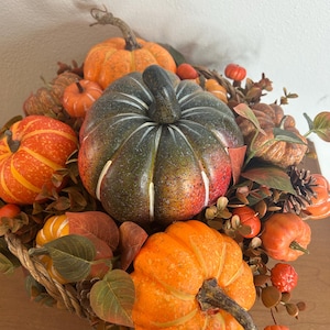May include: A decorative arrangement of pumpkins in various sizes and colors, including orange, green, and red. The pumpkins are surrounded by fall foliage, creating a festive autumn display. The arrangement is set in a woven basket.
