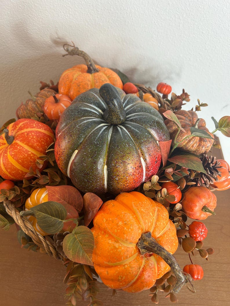 May include: A decorative arrangement of pumpkins in various sizes and colors, including orange, green, and red. The pumpkins are surrounded by fall foliage, creating a festive autumn display. The arrangement is set in a woven basket.