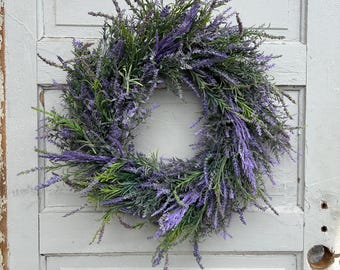 May include: A circular wreath made of faux lavender and greenery. The wreath features vibrant purple lavender blooms and green foliage, creating a natural and decorative accent. The wreath is displayed against a weathered, light gray door.