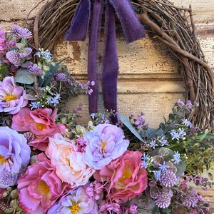 May include: A grapevine wreath adorned with a variety of faux flowers in shades of pink, purple, and peach. A deep purple velvet ribbon is tied at the top. The wreath is a decorative piece for the home.
