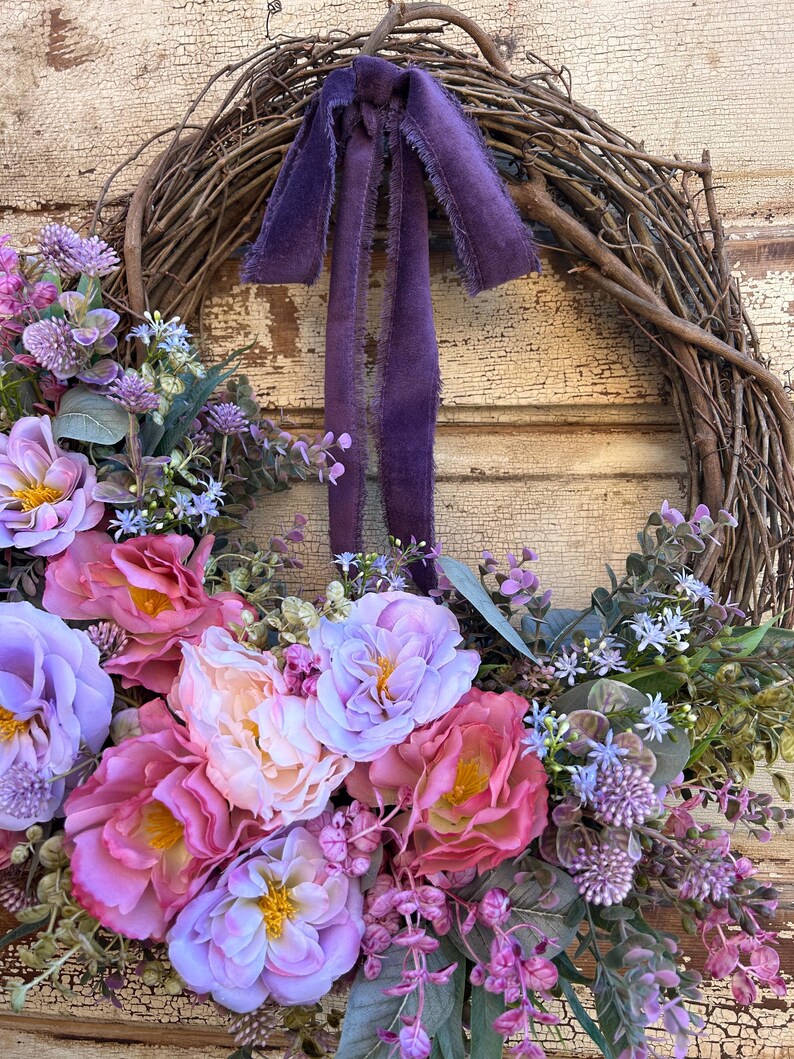 May include: A grapevine wreath adorned with a variety of faux flowers in shades of pink, purple, and peach. A deep purple velvet ribbon is tied at the top. The wreath is a decorative piece for the home.