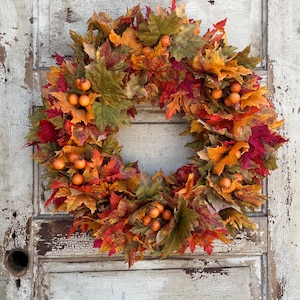 May include: A vibrant autumn wreath featuring a mix of red, orange, and green leaves, accented with small orange berries. The wreath is set against a distressed, white-painted wooden door, giving a rustic feel.