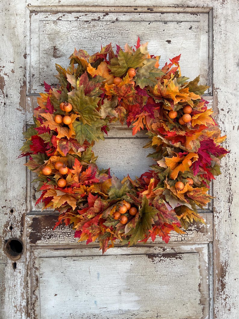 May include: A vibrant autumn wreath featuring a mix of red, orange, and green leaves, accented with small orange berries. The wreath is set against a distressed, white-painted wooden door, giving a rustic feel.