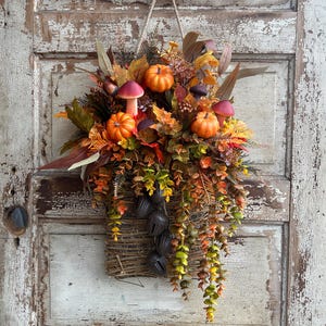 May include: A fall-themed wall hanging featuring a woven basket overflowing with autumn foliage, miniature orange pumpkins, and decorative mushrooms. The arrangement includes various leaves in shades of red, orange, and green, with hanging greenery and dark bells.