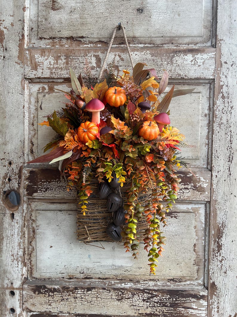 May include: A fall-themed wall hanging featuring a woven basket overflowing with autumn foliage, miniature orange pumpkins, and decorative mushrooms. The arrangement includes various leaves in shades of red, orange, and green, with hanging greenery and dark bells.