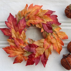 May include: A fall-themed wreath made of colorful artificial leaves in shades of red, orange, and yellow. Two small decorative pumpkins are also visible. The wreath and pumpkins are arranged on a striped, neutral-colored surface.
