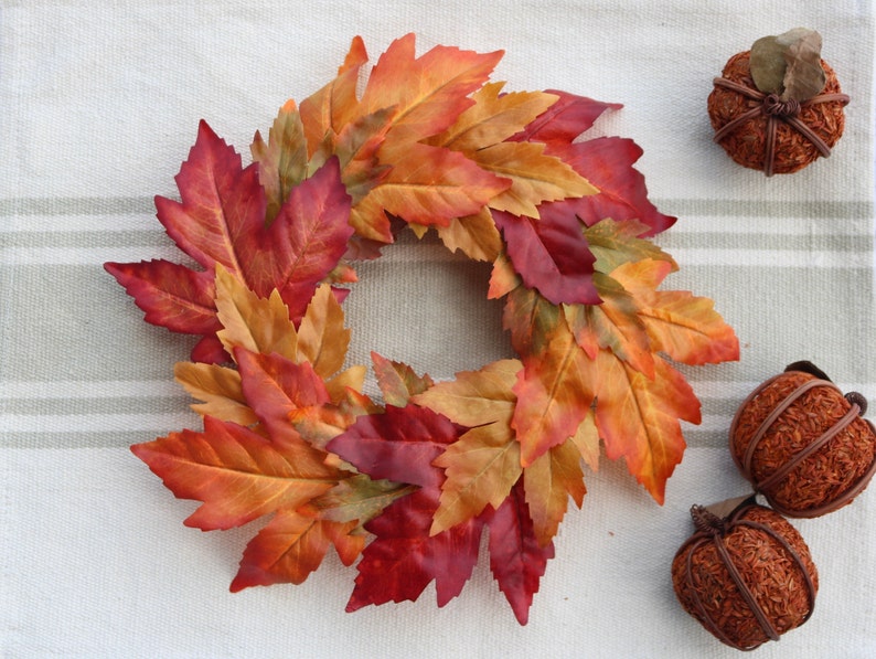 May include: A circular wreath of autumnal leaves in red, orange, and yellow hues. Two small decorative pumpkins are also present. The wreath and pumpkins are displayed on a striped, neutral-toned fabric surface.