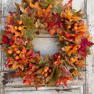 May include: A circular autumn wreath made of red, orange, and green leaves. Small orange berries are dotted throughout the wreath. The wreath is set against a weathered, white-painted wooden door, creating a rustic aesthetic.