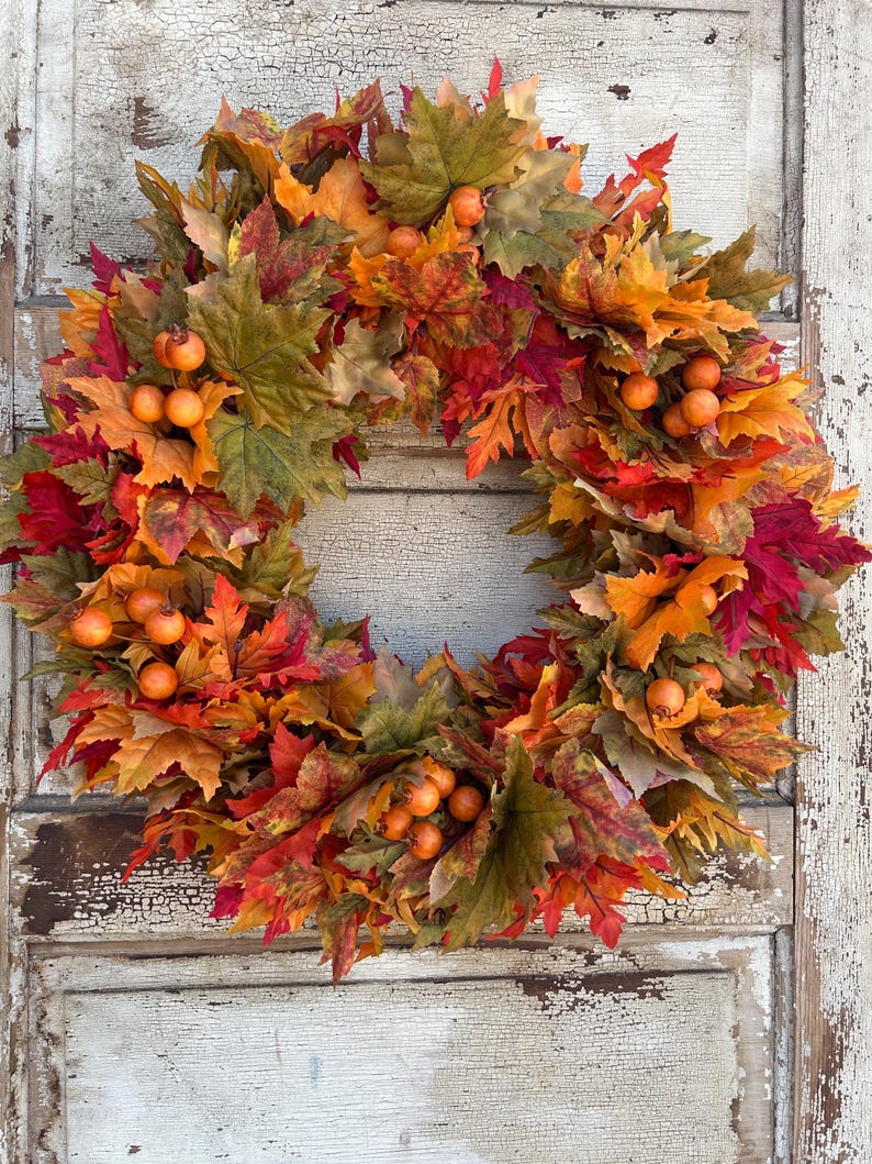 May include: A circular autumn wreath made of red, orange, and green leaves. Small orange berries are dotted throughout the wreath. The wreath is set against a weathered, white-painted wooden door, creating a rustic aesthetic.
