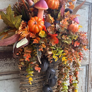 May include: A fall-themed decorative basket overflowing with faux pumpkins, mushrooms, and colorful foliage. The arrangement includes orange pumpkins, a pink mushroom, and various leaves in shades of red, orange, and green. Brown acorn-shaped bells hang from the basket.