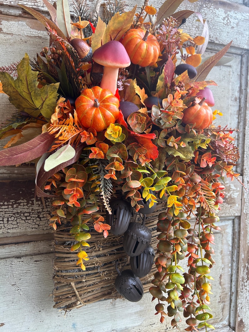 May include: A fall-themed decorative basket overflowing with faux pumpkins, mushrooms, and colorful foliage. The arrangement includes orange pumpkins, a pink mushroom, and various leaves in shades of red, orange, and green. Brown acorn-shaped bells hang from the basket.